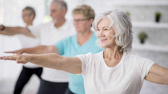 A multi-ethnic group of adult men and women are indoors in a fitness studio. They are wearing casual clothing while at a yoga class. A senior Caucasian woman is smiling while stretching out her arms.