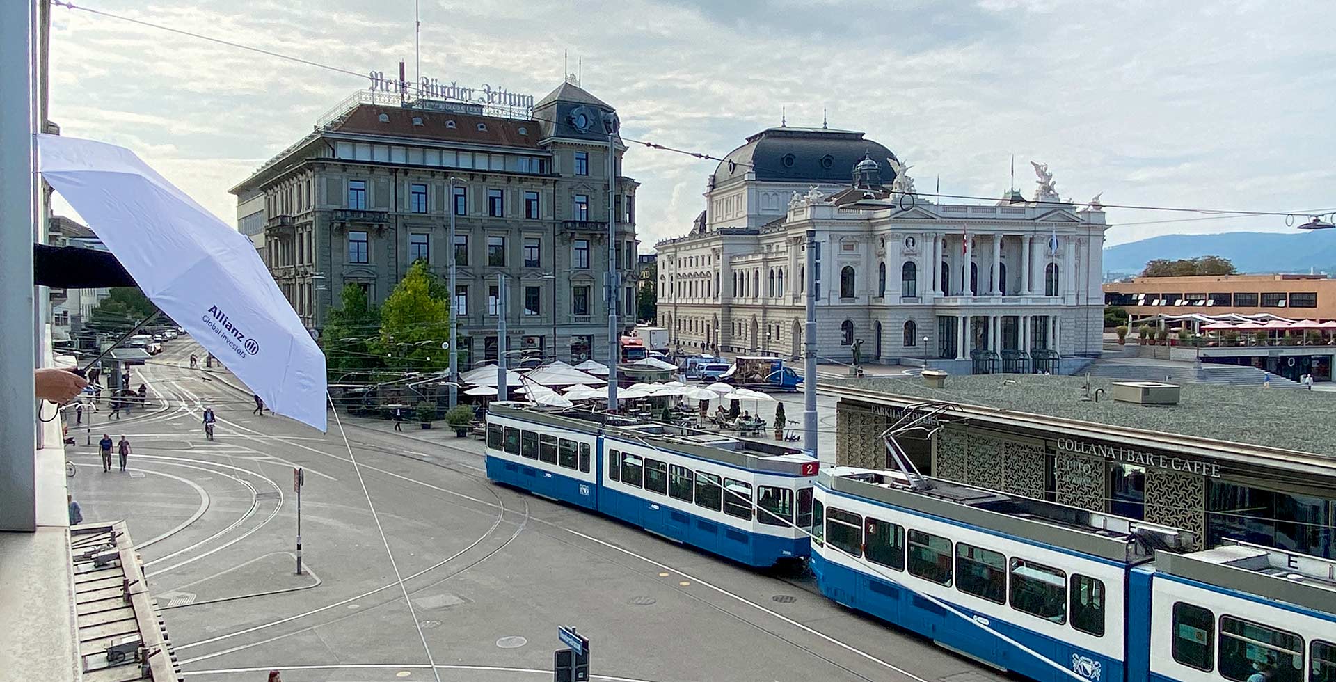 Eine Ansicht eines Stadtplatzes in Zürich mit vorbeifahrenden Straßenbahnen, Spaziergängern und einem Schirm mit dem Allianz-Logo im Vordergrund.